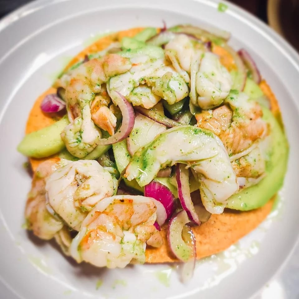 Mexican shrimp ceviche tostada with avocado, red onion, and green sauce on a white plate.