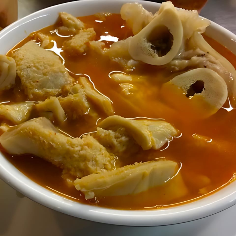 Close-up of a white bowl filled with Cocina Doña Chuyita's menudo, featuring tripe and bone marrow in a rich red broth.