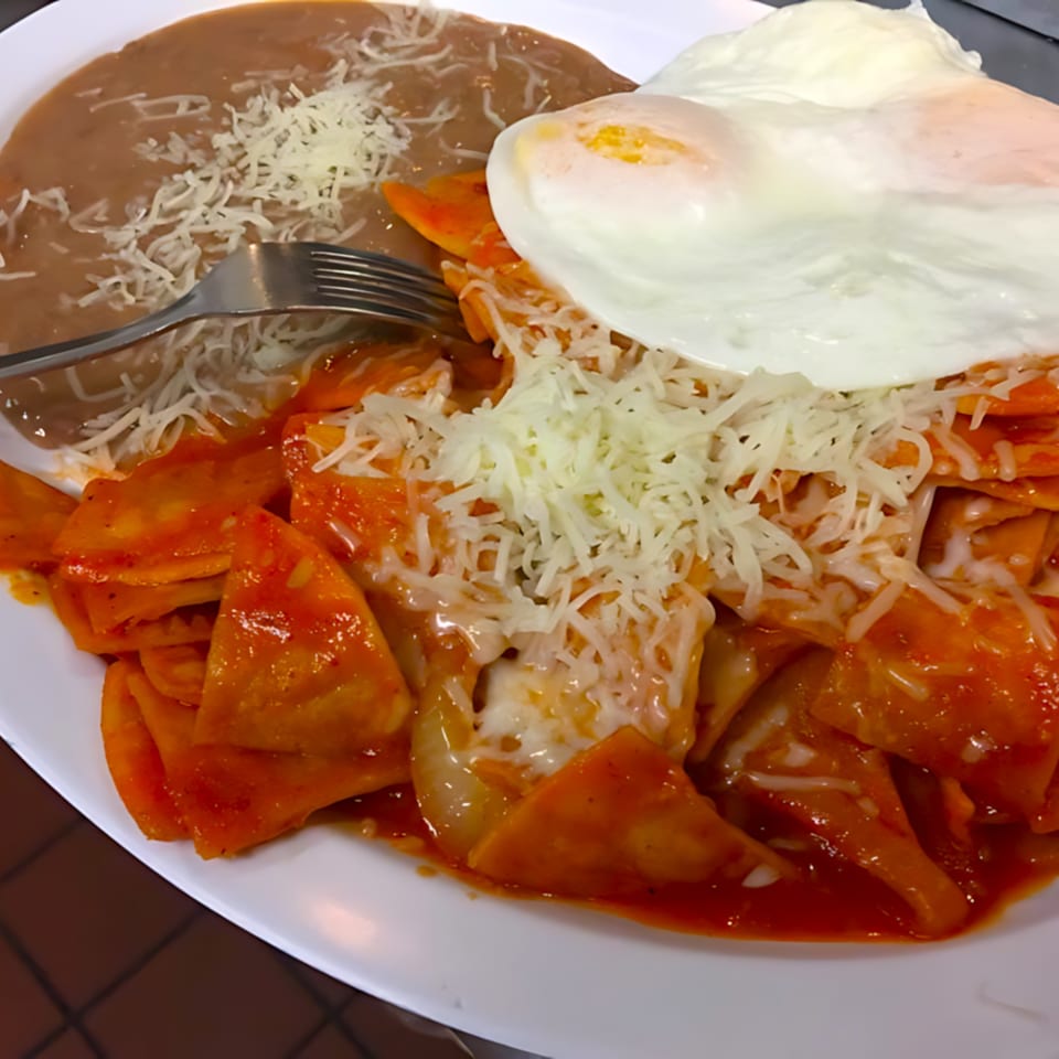 Chilaquiles rojos with two fried eggs, refried beans, and shredded cheese on a white plate.